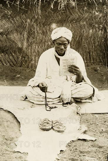 Nigerian man praying. A Nigerian man sits cross-legged on a mat, holding prayer beads in one hand whilst reading a page of scripture. An animal hide is spread out in front of him, on which are positioned a pair of shoes. Nigeria, circa 1915. Nigeria, Western Africa, Africa.