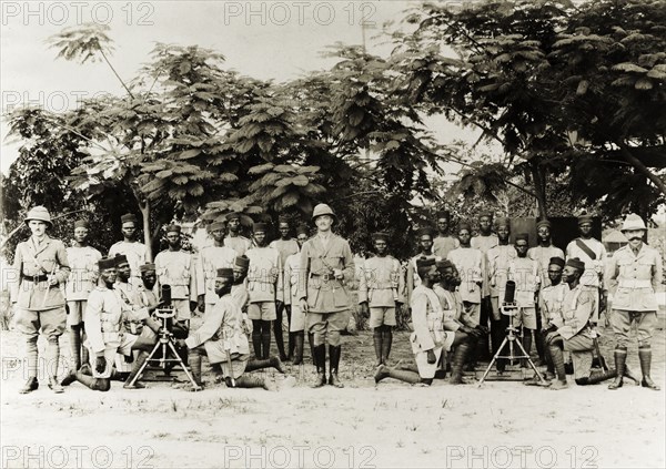 Artillery company of the Nigeria Regiment. Three British officers pose with an artillery company of the Nigeria Regiment of the Royal West African Frontier Force. Several African soldiers kneel around two machine guns on tripods at the front of the group. Nigeria, circa 1918. Nigeria, Western Africa, Africa.