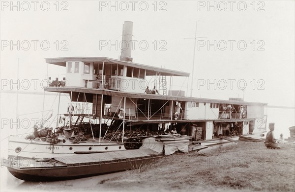 A paddle steamer named 'Sarota', Nigeria. A paddle steamer named 'Sarota' is moored to a riverbank in Nigeria. Several passengers are visible onboard, waiting for the vessel to leave shore. Nigeria, circa 1910. Nigeria, Western Africa, Africa.