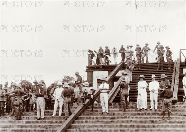 British colonial officers in solatopi hats. A group of British colonial officers, dressed in solatopi hats, gather outdoors on a flight of steps to watch something out of shot, perhaps a sporting event. Probably Nigeria, circa 1915. Nigeria, Western Africa, Africa.