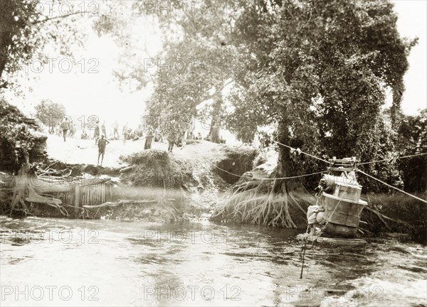 Transporting luggage across a river. A pile of luggage is stacked onto a raft, ready to be hauled across a river using a rope and pulley system. German East Africa (Tanzania), circa 1910. Tanzania, Eastern Africa, Africa.