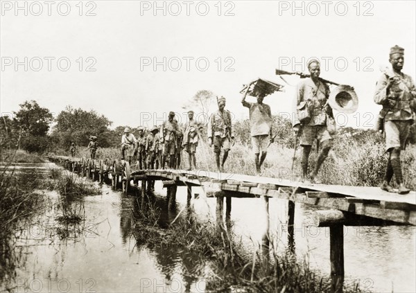 African schutztruppes on the march. A regiment of African schutztruppes march in single file across a wooden bridge over a river in German East Africa. German East Africa (Tanzania), circa 1910. Tanzania, Eastern Africa, Africa.