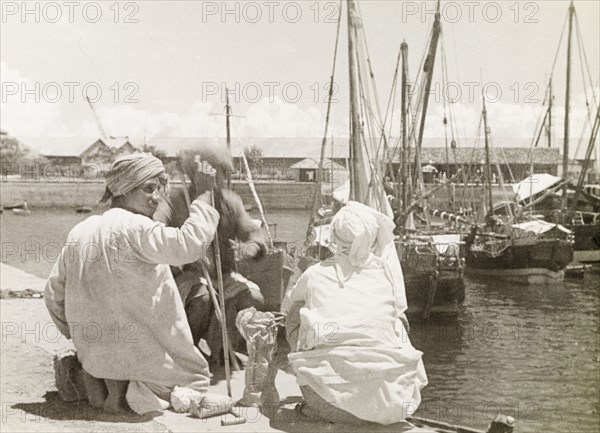 The Old Harbour at Stone Town. Three men sit beside the Old Harbour at Stone Town, with several dhows moored nearby. This harbour was once a centre of trade between East Africa and the Middle East until colonisation of the mainland in the 1880s transferred trade to Mombasa and Dar es Salaam. Stone Town, Zanzibar (Unguja, Tanzania), 1947. Stone Town, Zanzibar Urban/West, Tanzania, Eastern Africa, Africa.