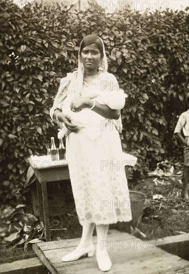 Indo-Trinidadian woman with baby. Portrait of an Indo-Trinidadian woman holding a baby in the crook of her arm. She wears a Western-style dress and shoes, with a traditional Indian 'dupatta' (head scarf). Siparia, Trinidad, circa 1931. Siparia, Trinidad and Tobago, Trinidad and Tobago, Caribbean, North America .