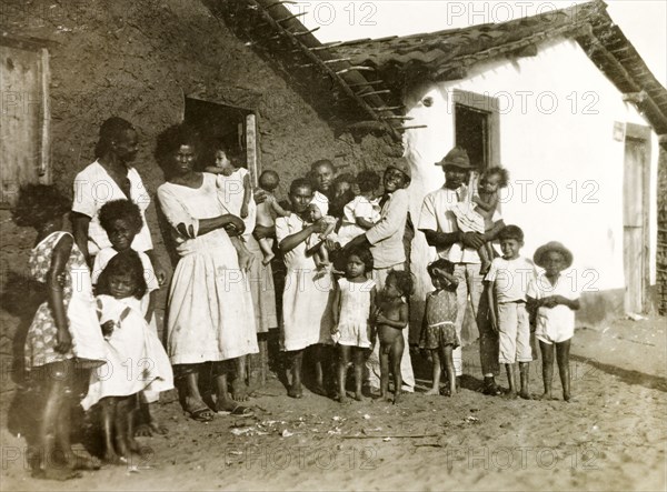 Brazilian families in Natal. A number of Brazilian families assemble for an informal portrait outside a row of single-storey houses. The outer wall of the nearest house has not yet been plastered, revealing its mud brick construction below. Natal, Brazil, circa 1931. Natal, Brazil, Brazil, South America, South America .