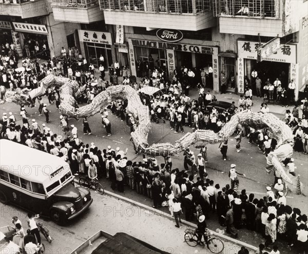 Dragon dance for the Tin Hau Festival. A Chinese dragon dance winds its way through crowds of spectators on a street in Yuen Long during celebrations for the Taoist Tin Hau Festival. An original caption comments: "Shop owners and schoolboys, housewives and factory workers all stop what they are doing: even the bus has to pull up on one side of the street to watch the parade". New Territories, Hong Kong, China, 1963., Hong Kong, China, People's Republic of, Eastern Asia, Asia.