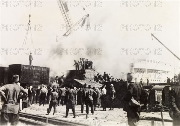 Fire at Jaffa Harbour. Smoke billows from the quayside at Jaffa Harbour as a team of men try to extinguish a fire with a hosepipe. Pictured during the Great Uprising (1936-39), this may have been an act of sabotage intended to damage infrastructure. Jaffa, British Mandate of Palestine (Israel), circa 1938. Jaffa, Tel Aviv, Israel, Middle East, Asia.
