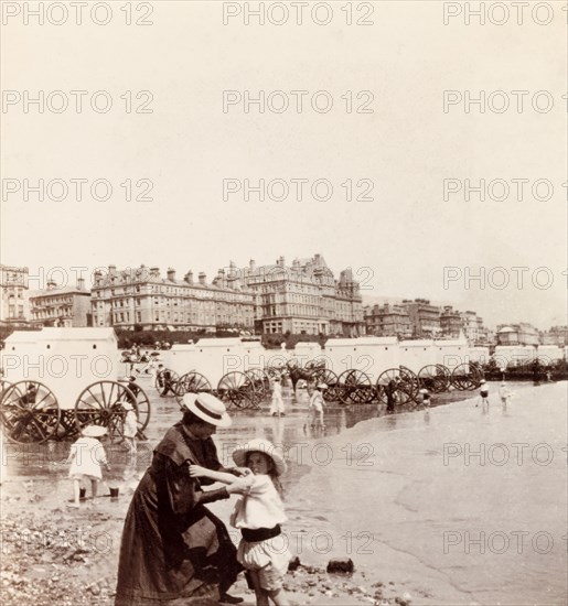 Bathing machines on Eastbourne beach. Minnie Murray rolls up her daughter's sleeves on the beach at Eastbourne. Behind them on the sand are a number of bathing machines, designed to give bathers modest access to the water. Eastbourne, England, 1907. Eastbourne, Sussex, England (United Kingdom), Western Europe, Europe .