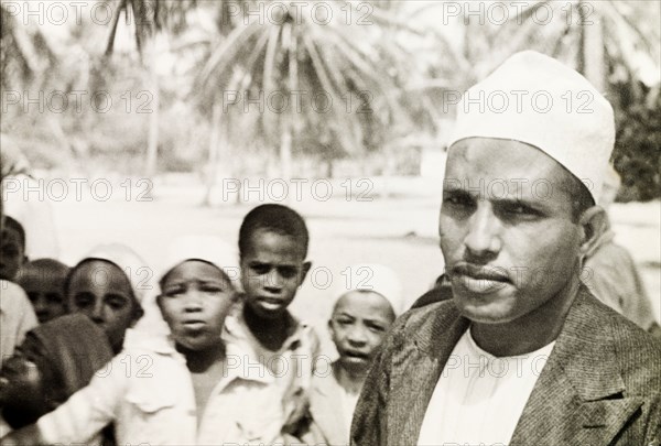 A Surahi type'. A Muslim man (possibly a teacher) stands in front of a group of children, staring into the camera. He wears a white kufi (cap) and is identified in a related caption as a 'Surahi type'. Possibly Mombasa, Kenya, 1947. Mombasa, Coast, Kenya, Eastern Africa, Africa.