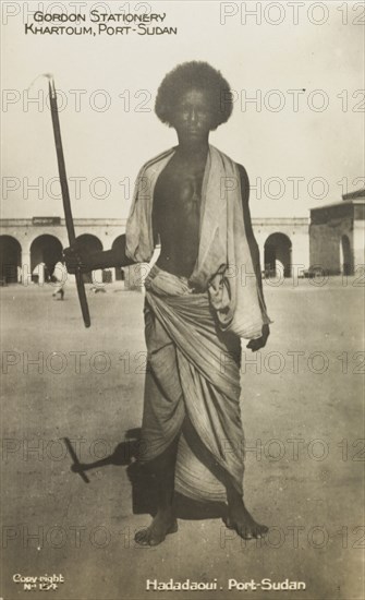 Portrait of a Hadadaoui man. Full-length portrait of a Hadadaoui man, standing barefoot in a courtyard holding a hooked stick. Khartoum, Sudan, circa 1927. Khartoum, Khartoum, Sudan, Eastern Africa, Africa.