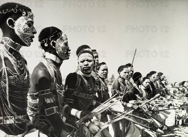 Tanganyikan dancers in ceremonial attire. Official publicity shot for the Tanganyikan government showing a line of male dancers in ceremonial costume. Wearing beaded jewellery including necklaces, armbands and chest-pieces, their faces are vividly painted with white dots. They dance holding clubs and bundles of short, straight sticks. Tanganyika Territory (Tanzania), circa 1960. Tanzania, Eastern Africa, Africa.