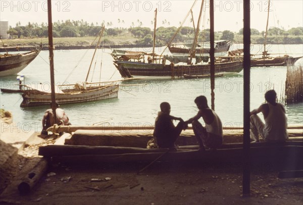 Dhows in Kilindini harbour'. Three men sit in the shade at Kilindini harbour, watching the dhows as they enter and leave the port. Mombasa, Kenya, April 1964. Mombasa, Coast, Kenya, Eastern Africa, Africa.