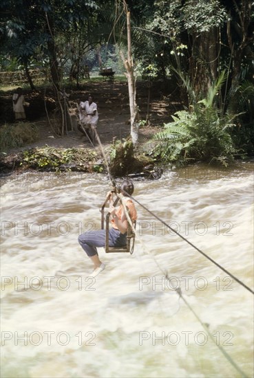 Elisabeth Lang Brown crosses to Kyantsore Island. Elisabeth, the wife of District Forestry Officer James Lang Brown, crosses the rapids of Kagera River by way of a cable chair lift. A related caption comments that she was crossing to Kyantsore Island to visit the Countess Antoinette Nuti (Toni), a colonial settler who had taken a lease on the island from the Omukama (King) on Ankole. Ankole, West Uganda, November 1963., West (Uganda), Uganda, Eastern Africa, Africa.