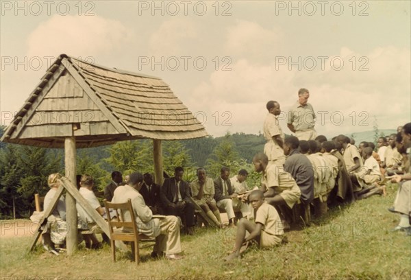 Open day at Mafuga Forest nursery. George Webster (Chief Conservator of Forests) addresses a crowd of Ugandan chiefs, villagers and schoolchildren during a forestry open day at Mafuga. To his right stands Forest Ranger Bitarabeho, who translates into Rukiga. A related image identifies the structure on the left as a bus stop constructed by local forestry workers. Mafuga, Kigezi, South West Uganda, September 1963., West (Uganda), Uganda, Eastern Africa, Africa.