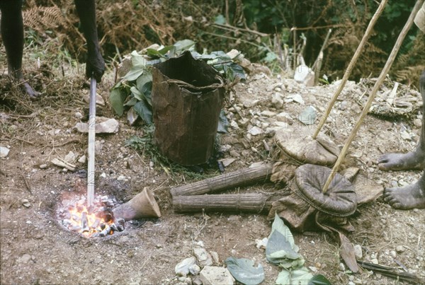 Iron Age forge in Mafuga Forest. A forge used by forestry workers to harden the tips of rock drills whilst clearing a track to Mafuga Forest nursery. A related caption explains: "A charcoal fire... was fanned by two... clay pipes and bellows made of a goat-skin... The white hot tips of the drills were hammered to a chisel edge, reheated and tempered in a battered debbi of water". Kigezi, South West Uganda, September 1963., West (Uganda), Uganda, Eastern Africa, Africa.