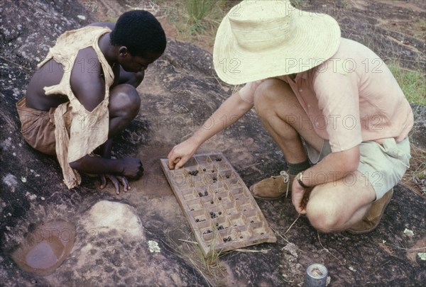 Playing omweso at Kojja. A British man identified as Martin Beadle squats down to play omweso ('mweso'), an ancient mancala game, with a Ugandan herd boy. A related caption comments: "(The game) is normally played on a portable wooden board with wild banana seeds as 'men'". Kojja, West Uganda, 1963., West (Uganda), Uganda, Eastern Africa, Africa.