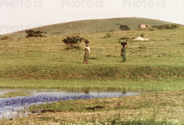 Two women collecting water. Two women balance containers on their heads as they collect water from a pool at Bubdare dam. Mbarara, West Uganda, September 1963., West (Uganda), Uganda, Eastern Africa, Africa.