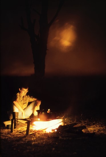 Camp fire at Ishasha River Camp. Elisabeth, the wife of District Forest Officer James Lang Brown, cooks supper on a camp fire at Ishasha River Camp in the Queen Elizabeth National Park. According to a related caption, the faint red glow emanating from the horizon behind her comes from a bush fire raging in the Congo across the border. West Uganda, July 1962., West (Uganda), Uganda, Eastern Africa, Africa.
