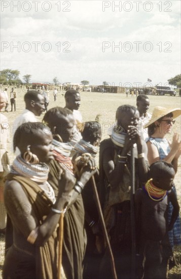 An intrigued audience for the Uganda Police Band'. A group of Pian women and children watch a performance by the Uganda Police Band at the Nabilatuk County Show. To their left stands Elisabeth Lang Brown, the wife of District Forestry Officer James Lang Brown, and behind, a British man identified as Charles Lamb. Nabilatuk, North East Uganda, October 1959., East (Uganda), Uganda, Eastern Africa, Africa.