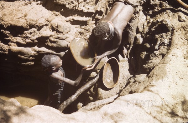 Waterhole in Mbaru. A young Suk (Pokot) woman stoops to collect a gourd full of water, passed to her by a small girl from the bottom of a deep waterhole. Mbaru, North East Uganda, 1959., North (Uganda), Uganda, Eastern Africa, Africa.