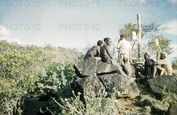 Henry Osmaston takes a bearing'. Accompanied by Suk porters, Henry Osmaston (Working Plans Officer of the Forest Department) stands at the base of a 'kituti' (cairn) marked with bamboo sticks, surveying Pcholio Peak in the Karasuk hills. Part of a forestry survey team, he uses a theodolite to locate and measure the distance to the next 'kituti'. Kenya, May 1959., East (Uganda), Uganda, Eastern Africa, Africa.