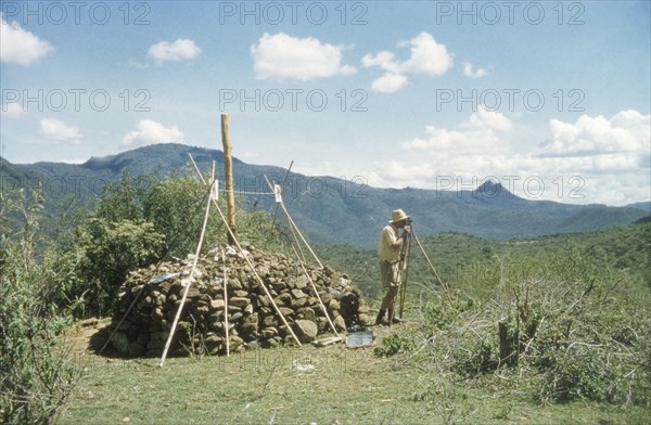 Henry Osmaston surveys Pcholio Peak. Henry Osmaston (Working Plans Officer of the Uganda Forest Department) stands at the base of a 'kituti' (cairn) marked with bamboo sticks, surveying Pcholio Peak in the Karasuk hills. Part of a forestry survey team, he uses a theodolite to locate and measure the distance to the next 'kituti'. Kenya, May 1959., East (Uganda), Uganda, Eastern Africa, Africa.