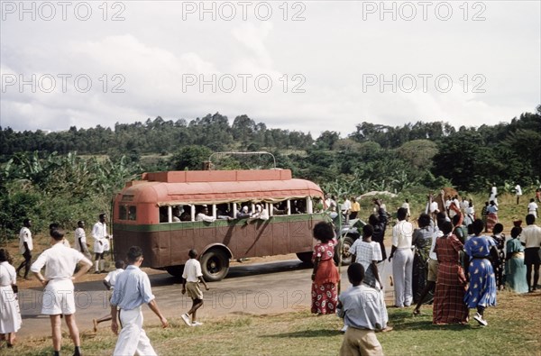 The faithful'. Crowds of people wave to a bus on a rural road during celebrations for the return from exile of Edward Mutesa II, Kabaka (King) of Buganda. An original caption comments: "The faithful - the women did not cut their hair during the exile of their King". Near Lake Victoria, Central Uganda, October 1955., Central (Uganda), Uganda, Eastern Africa, Africa.