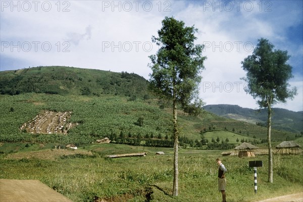 A village in the Belgian Congo. A British District Officer stands on the road beside a village in the Belgian Congo. An original caption comments on "the tidiness in contrast to Ugandan villages, where each hut is surrounded by its own shamba". Belgian Congo (Democratic Republic of Congo), 1956. Congo, Democratic Republic of, Central Africa, Africa.