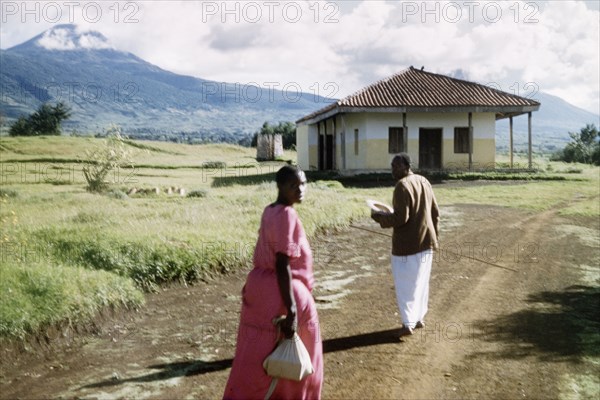 A Gombolola chief and his wife. A Gombolola chief and his wife walk towards his office below Mount Muhabura (Muhavura). An original caption comments: "The Gombolola was a middle ranking community, comprising a number of tiny villages or murukas, each with its own junior chief. These divisions were the bedrock of indirect rule". Kigezi, South West Uganda, 1956., West (Uganda), Uganda, Eastern Africa, Africa.