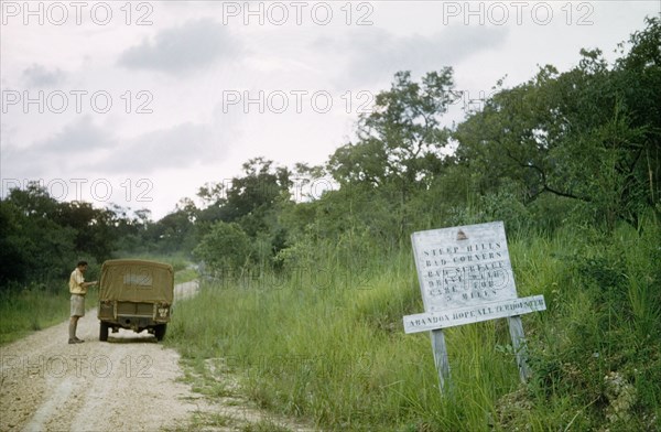 Abandon hope all ye who enter here'. Henry Osmaston (District Forestry Officer of the West Nile district) checks his jeep on the road to Arua. The sign in the foreground reads: "Steep hills, bad corners, bad surface, drive with care for 3 miles. Abandon hope all ye who enter here". North Uganda, 1956., North (Uganda), Uganda, Eastern Africa, Africa.