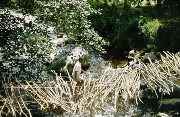 Ramshackle rope bridge in Kyarumba. A man crosses an old, ramshackle rope bridge above a river in Toro. The bridge was soon to be replaced, using local timber and unpaid labour, as part of a community development project supported by the District Forest Office. Kyarumba, West Uganda, 1956., West (Uganda), Uganda, Eastern Africa, Africa.