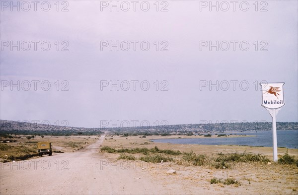 A rural road in Katwe. A Mobiloil sign stands beside a rural road in Katwe to Fort Portal, a fishing village located in the Queen Elizabeth National Park. Katwe's volcanic salt lake is visible in the distance. Katwe, West Uganda, August 1956., West (Uganda), Uganda, Eastern Africa, Africa.