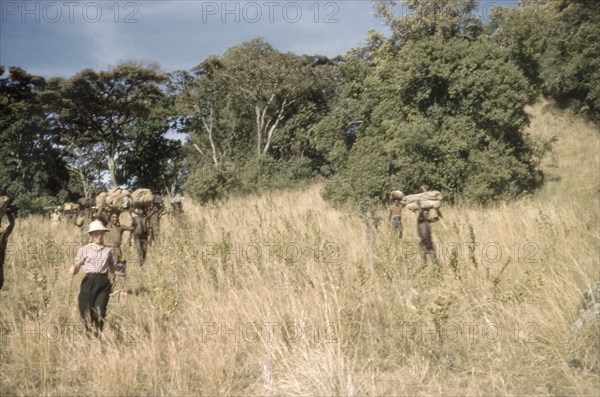 Coming down the mountain. Jenny Cleeve, the wife of Assistant District Commissioner John Cleeve, walking down a grassy slope on the descent from Mount Napak in Karamoja. Several Ugandan safari porters follow behind, laden with equipment belonging to a British forestry survey team. North East Uganda, December 1958., East (Uganda), Uganda, Eastern Africa, Africa.