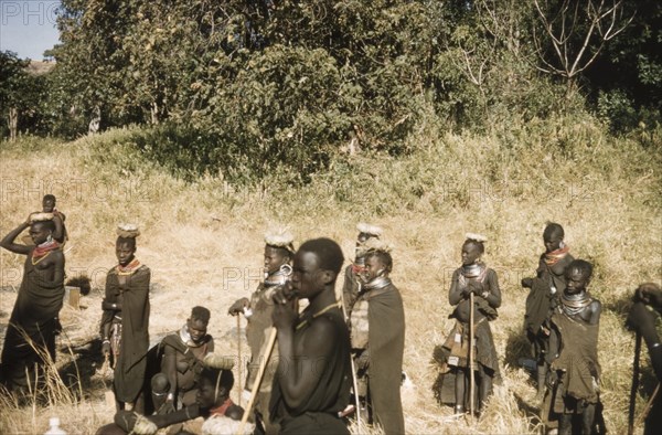 Porter's wives on a safari to Mount Napak. A group of porter's wives, wearing head pads and holdings staffs, look on with concern as their husbands dispute the weight of equipment to be carried on safari. The group were assisting a British forestry survey team on a trip to Mount Napak in Karamoja. North East Uganda, December 1958., East (Uganda), Uganda, Eastern Africa, Africa.