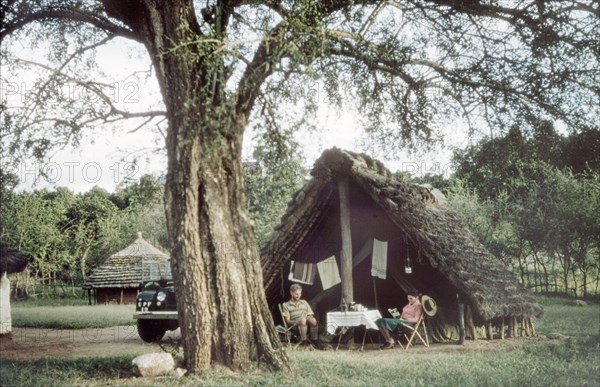 Kaabong rest camp. District Forest Officer James Lang Brown relaxes at a rest camp in Kaabong with his wife, Elisabeth, in front of the grass-thatched 'banda' which protected the tent from the midday sun. The tree in the foreground is a Desert Date (Balanites aegyptiaca). Kaabong, North East Uganda, 1958., North (Uganda), Uganda, Eastern Africa, Africa.