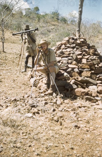 Surveying with 'kituti' markers. District Forest Officer James Lang Brown (right) is accompanied by Forest Ranger Langoya on a survey safari in the Karasuk hills on the Kenya-Uganda border. The men use a range finder and compass to measure the distance between one 'kituti' (cairn) marker and the next. A related caption comments: "When plotted on the map, the line of bituti... will form the legal boundary of a new forest reserve from which marauding goats will be excluded". Kenya, 1959., East (Uganda), Uganda, Eastern Africa, Africa.