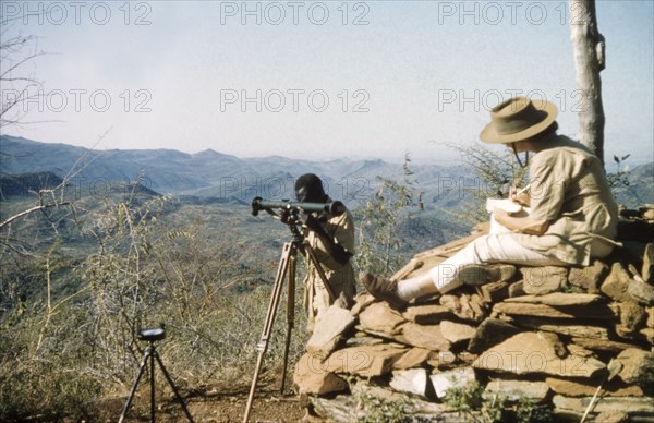 Elisabeth Lang Brown records survey data. Elisabeth, the wife of District Forest Officer James Lang Brown, sits on a 'kituti' (cairn) as she records data during a survey safari to the Karasuk hills on the Kenya-Uganda border. In front of her, a Forest Ranger uses a range finder to measure the distance to the next 'kituti'. A related caption comments: "When plotted on the map, the line of bituti cairns will form the legal boundary of a new forest reserve from which marauding goats will be excluded". Kenya, 1959., East (Uganda), Uganda, Eastern Africa, Africa.