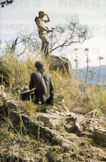 James Lang Brown looks for a 'kituti'. Accompanied by a Suk porter, District Forest Officer James Lang Brown holds a compass in one hand as he surveys the land for a 'kituti' (cairn) marker on a new forest reserve in the Karasuk hills. Kenya, January 1959., East (Uganda), Uganda, Eastern Africa, Africa.