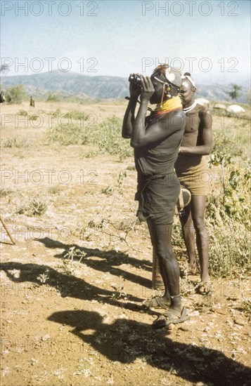 Lotuke with binoculars. Lotuke, a Suk porter working with a British forestry survey team, peers through binoculars to locate a 'kituti' (stone cairn) marker on the boundary of a new forest reserve in Karasuk on the Kenya-Uganda border. North East Uganda, January 1959., East (Uganda), Uganda, Eastern Africa, Africa.