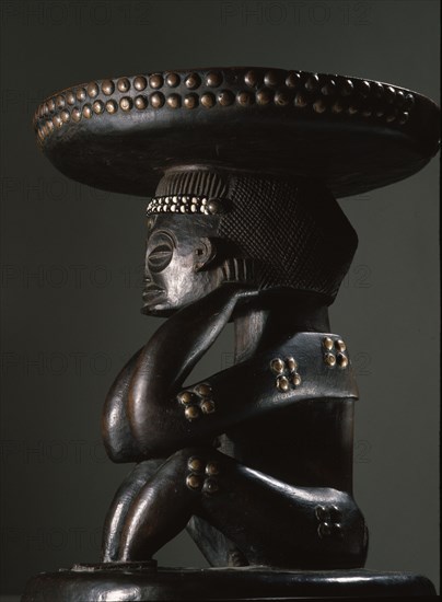 Caryatid stool supported by a depiction of a matrilineal ancestor, her seated posture with hands behind the ears indicates mourning, reminding the living of the necessity of sacrifices to the dead