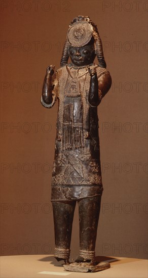 A large figure, one of the Tsoede bronzes, wearing an outer garment covered with cowrie shells, a necklace of leopards teeth and a pectoral with representations of a ram and birds