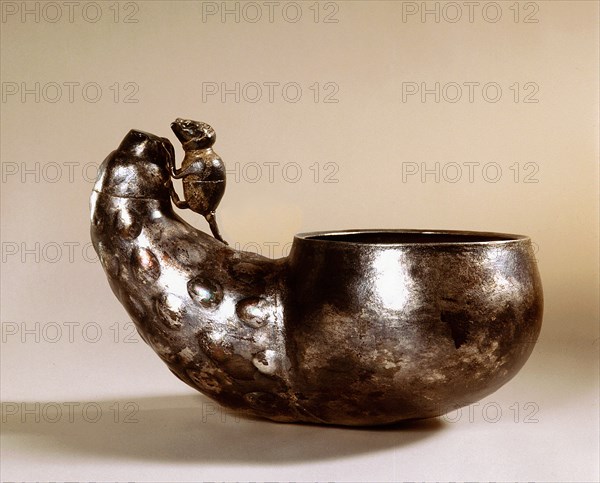 A cup made of beaten metal with a handle in the form of a mouse climbing over a vegetable
