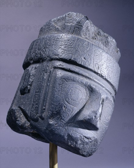 Head from a pillar statue with finely incised features and wearing a classic cornered hat decorated with a frieze of mythical feline figures