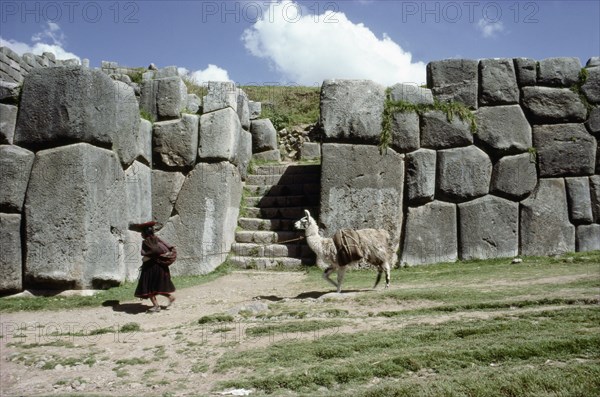 Quechua woman and llama walking past a monumental Inca doorway and wall at Sacsahuaman
