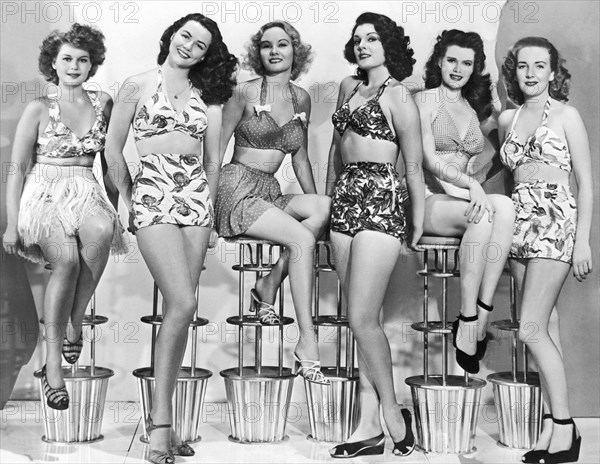 United States:  c. 1952.
Six attractive young women in two piece bathing suits sit in a row on modern stools.
