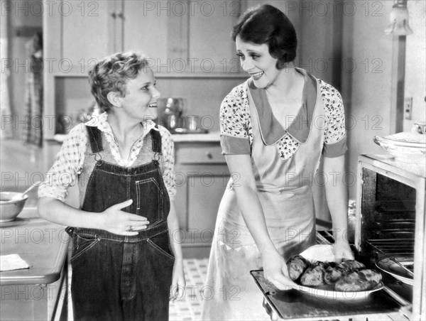 United States:  c. 1934.
A young teenager eagerly antiicipates the dinner his mother is taking out of the oven.