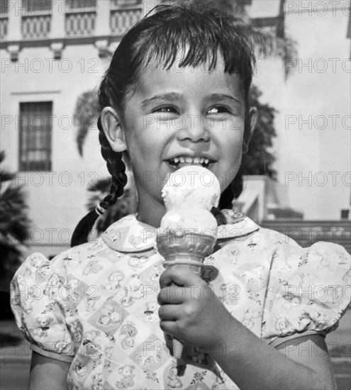California:  c. 1948.
A smiling young ethnic girl in pigtails enjoys eating a double dip ice cream cone on a warm summer day.