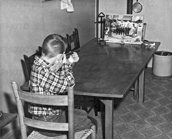 United States:  c. 1950.
A boy shoots at a birds on a wire game with a cork gun on the family table.