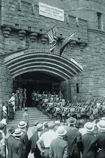 71st Street Armory in New York welcomes uniformed doughboys in formation with rifles and full pack; a sign above the portcullis of the emulated stone castle invites men to enlist to fight in World War I. 1917