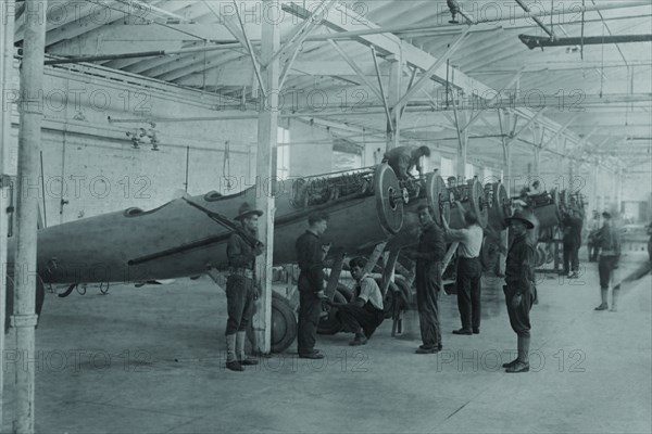 A line of airplanes is being assembled under the watchful eye of a gun-toting soldier as they are being completed to be sent to the European war; the factory is at College Point, Queens 1917